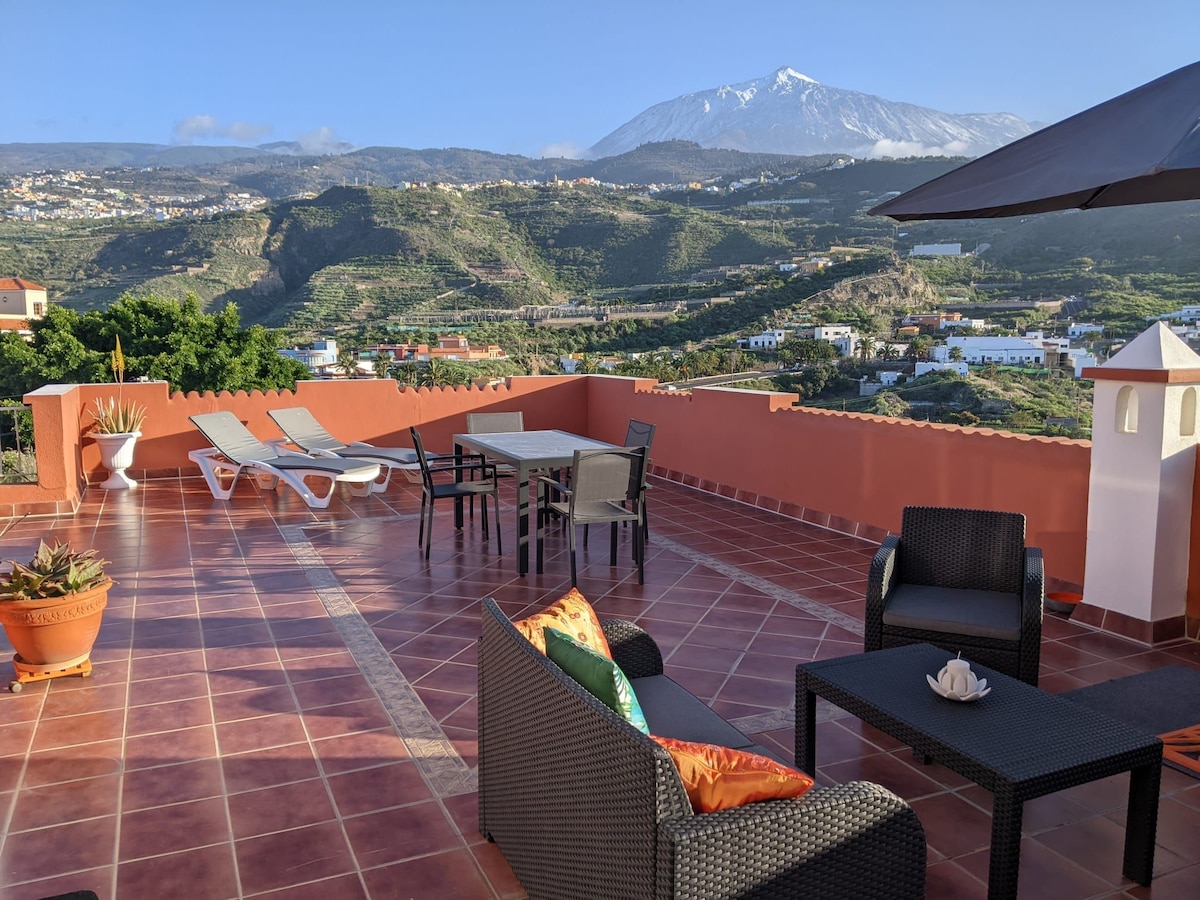 A spacious private terrace is shown, featuring outdoor seating with comfortable chairs and a dining table. Sun loungers are positioned nearby. The background reveals a mountain view, with lush greenery surrounding the area, enhancing the connection to nature.