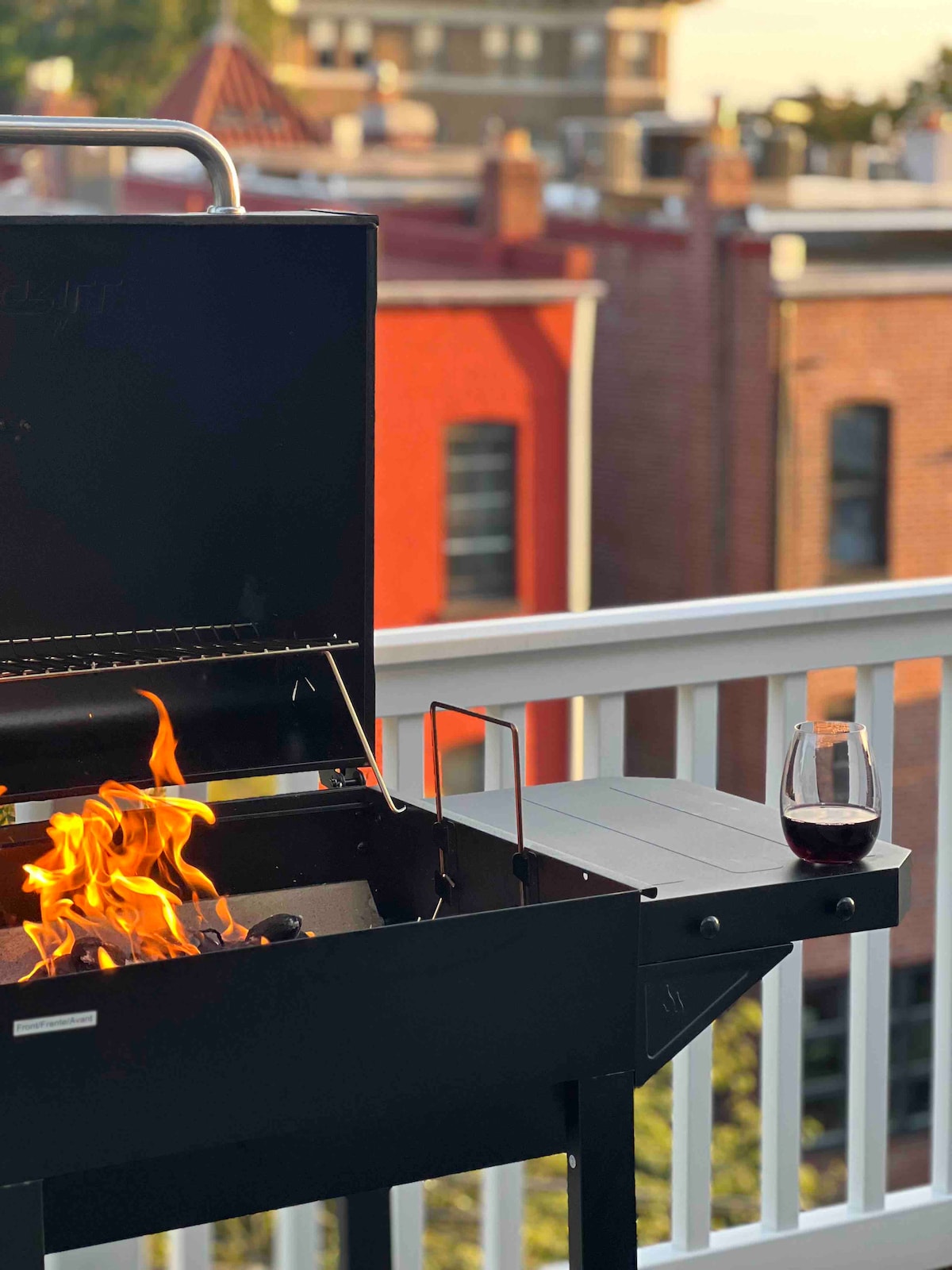A balcony grill is prominently featured, with flames flickering inside. A glass of red wine rests on the adjacent surface, providing an inviting setting for outdoor dining. Vibrant red and brick buildings are visible in the background, adding an urban touch.