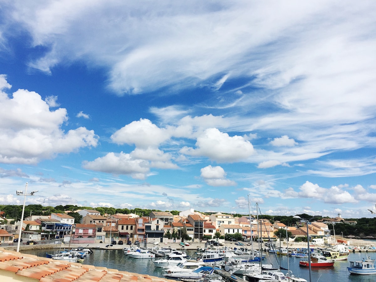 A view of the charming harbor is displayed, with several boats anchored in the water. Colorful buildings line the shore under a blue sky embellished with scattered clouds.