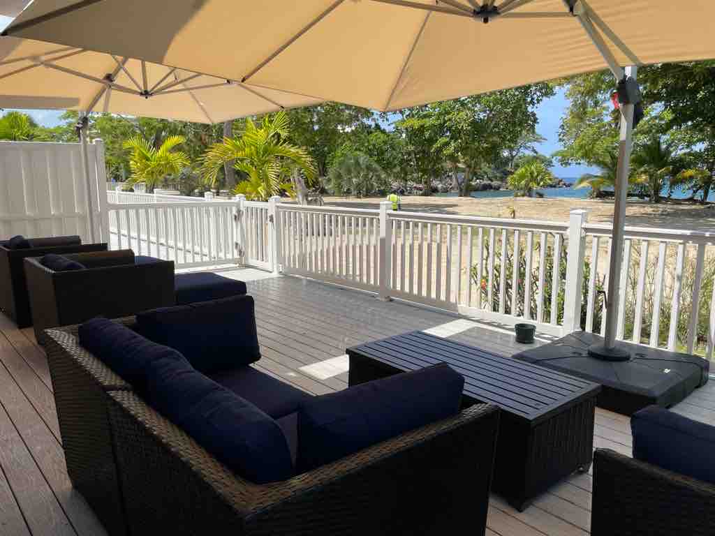 An outdoor patio area is seen with a set of wicker seating, featuring dark blue cushions. A wooden table is positioned in the center, and a beige umbrella provides shade. In the background, a view of palm trees and the beach is visible beyond the white railing.