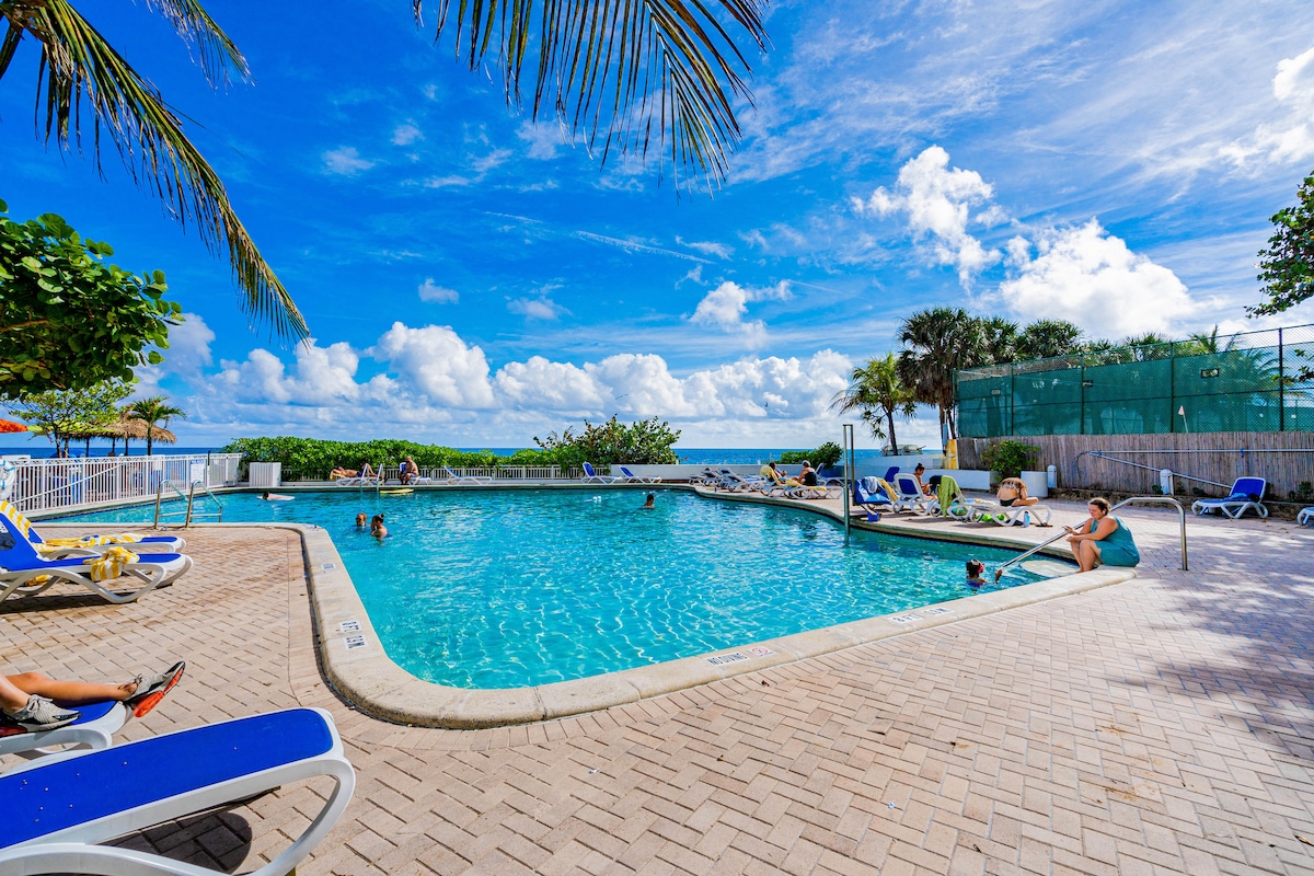 A large heated pool is surrounded by lounge chairs under a sunny sky. Lush palm trees and greenery frame the pool area, offering a relaxed atmosphere. The sparkling water reflects the clouds, inviting guests to enjoy leisure time by the pool.