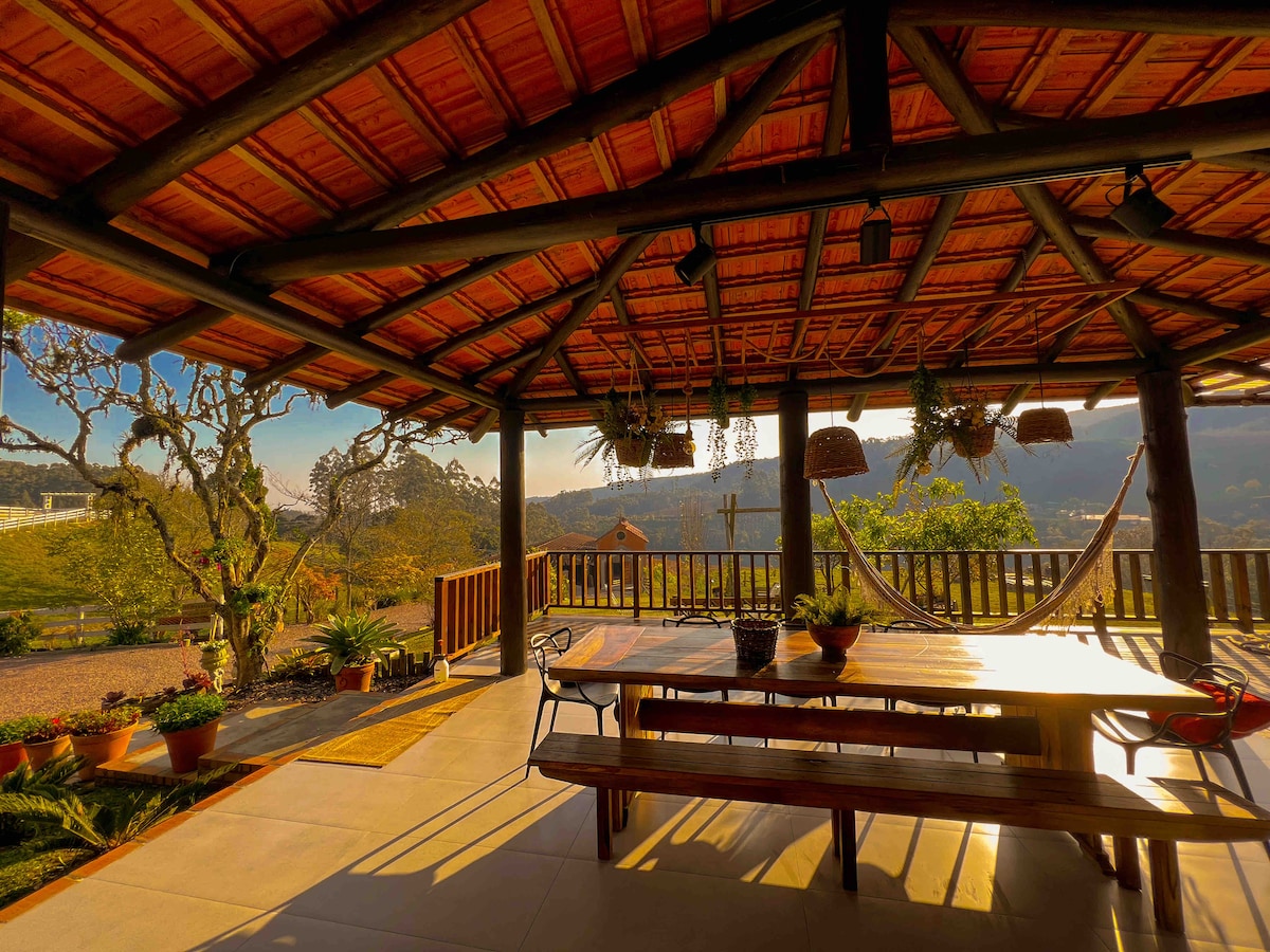 A covered outdoor dining area features a large wooden table surrounded by chairs. Hanging plants and lanterns adorn the ceiling, while a hammock is suspended nearby. The backdrop includes greenery and distant hills, with warm light highlighting the space.