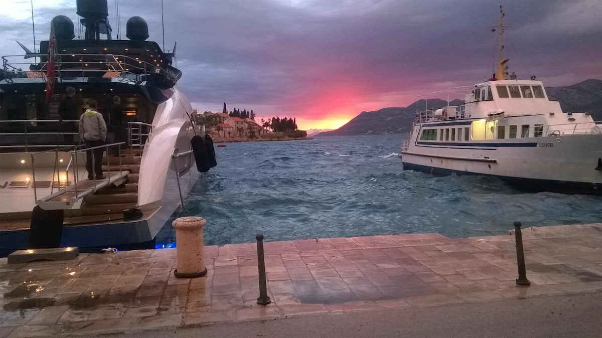 A view of the harbor showcases two boats, one large yacht and a smaller vessel, docked in the water. Colorful sunset hues blend in the sky, with vibrant reflections shimmering on the waves. The quayside path is partially visible, framed by stone posts.