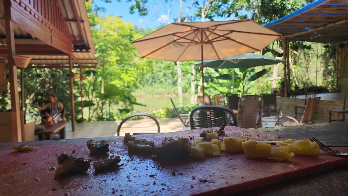 A rustic outdoor dining area is framed by lush greenery. A wooden table displays remnants of a meal, with partially eaten food. In the background, expansive umbrellas provide shade over seating arrangements, while glimpses of the river and surrounding foliage are visible.