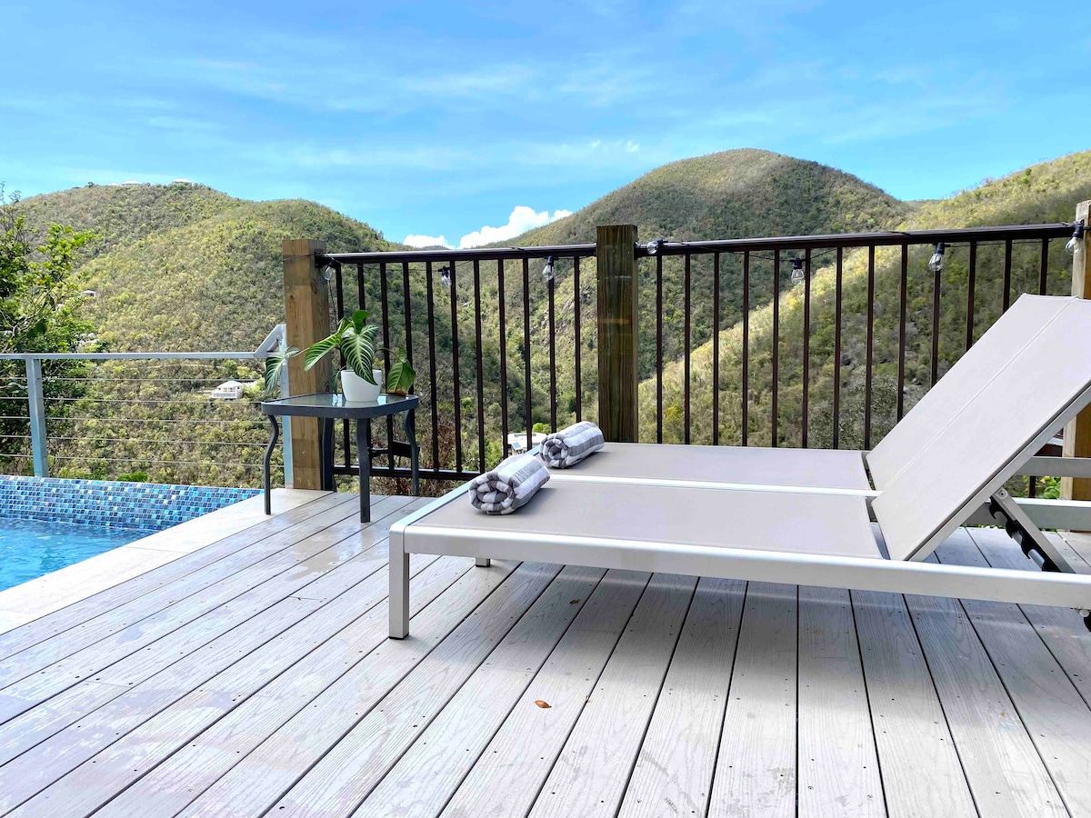 A lounging chair is positioned beside a sparkling blue pool, overlooking lush, rolling hills. A small table is nearby, holding a potted plant. Soft towels are neatly arranged on the chair, inviting relaxation in the serene outdoor space.