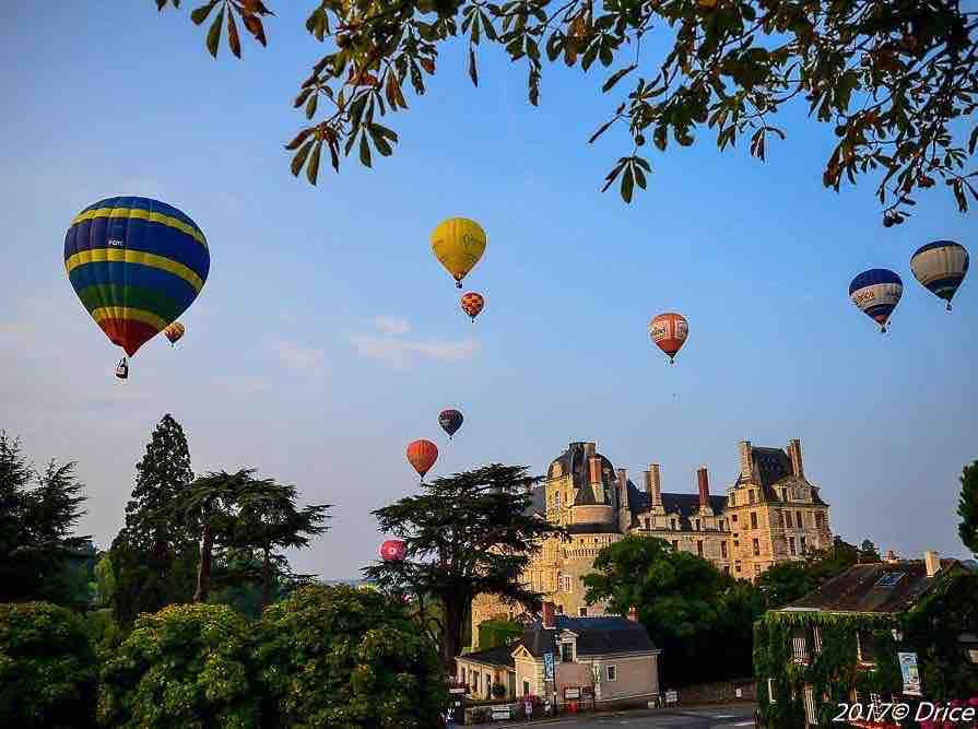 A vibrant sky is filled with numerous hot air balloons in various colors, soaring above a historical castle. The castle's architecture is visible amidst lush trees, with the scenery reflecting a serene, picturesque landscape. The scene suggests a tranquil atmosphere for outdoor exploration.