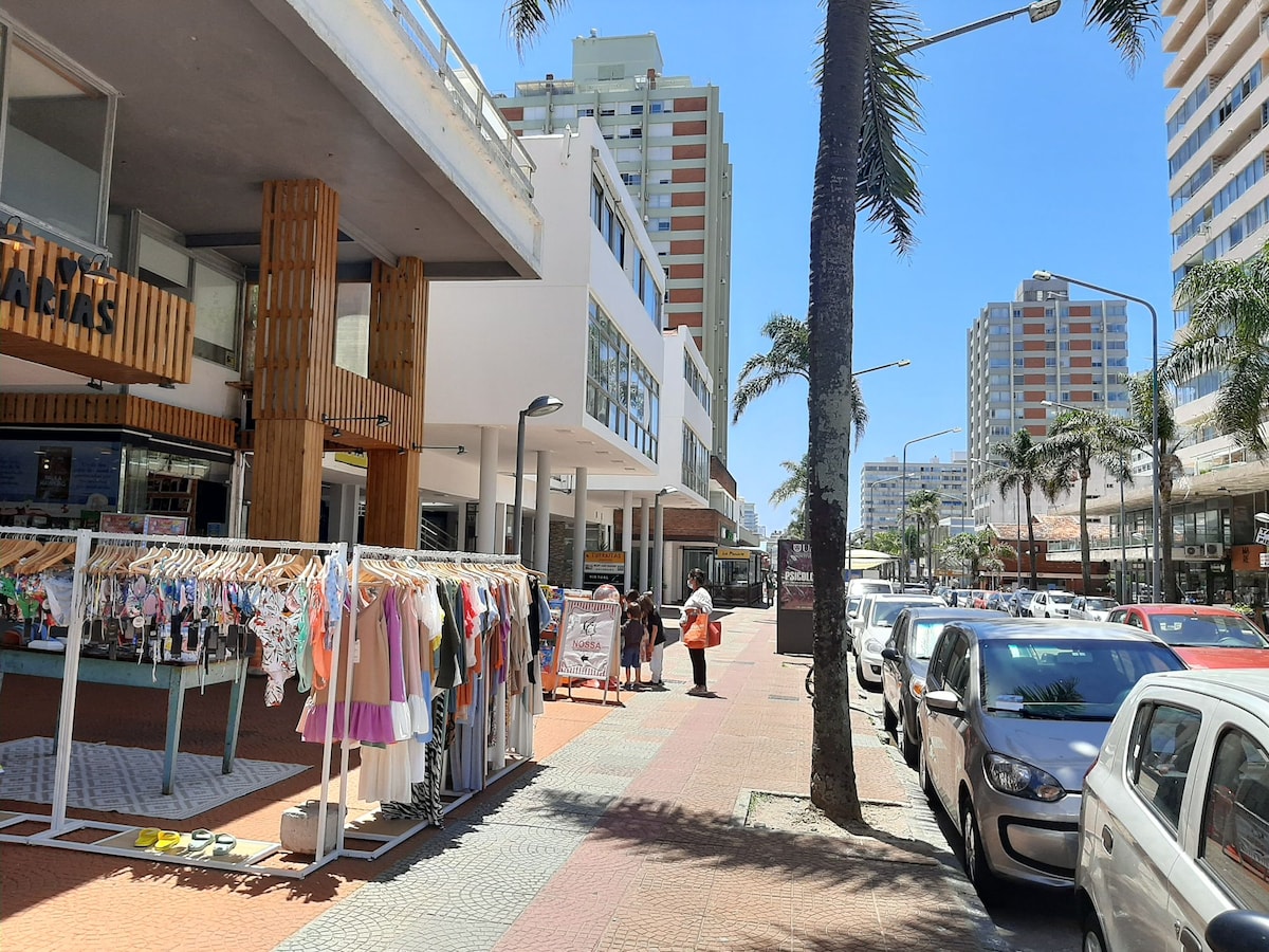 A vibrant street scene is depicted, featuring storefronts and a clothing rack overflowing with colorful garments. Palm trees provide shade to pedestrians walking along the neatly paved sidewalk. Modern high-rise buildings can be seen in the background, contributing to the lively atmosphere of the urban setting.