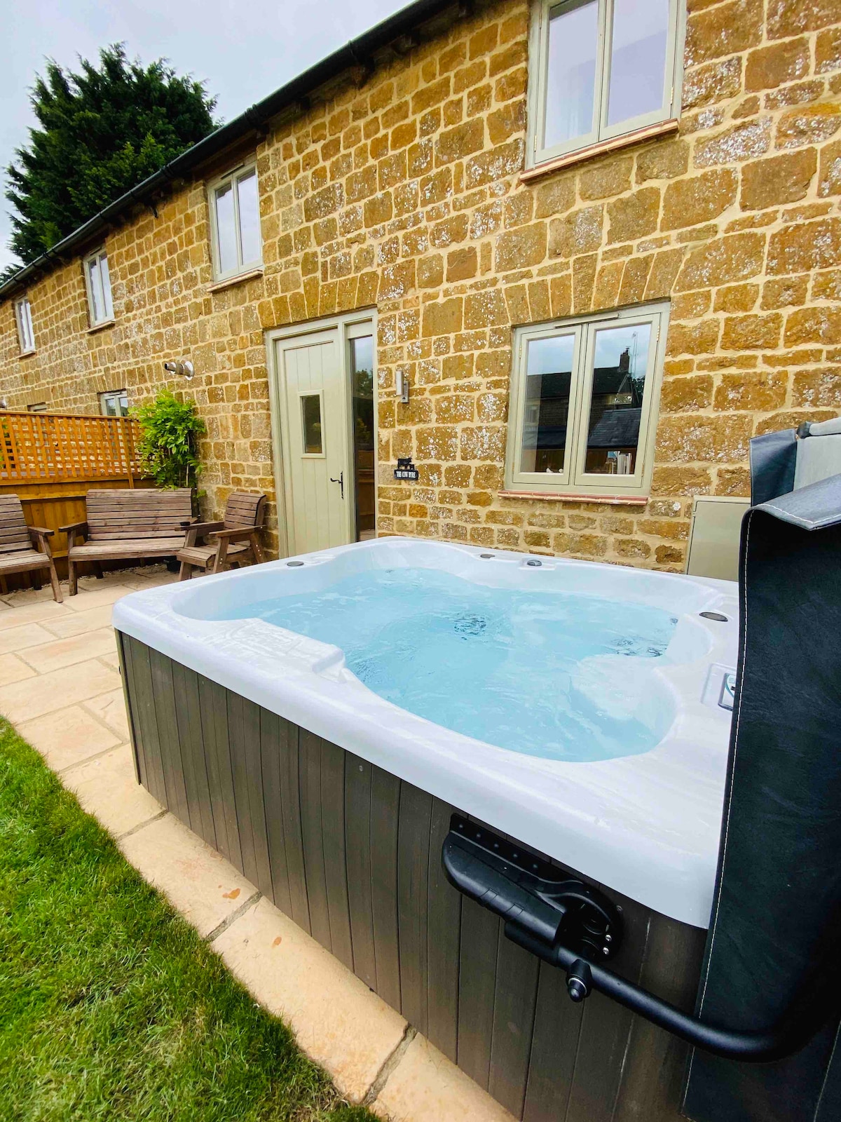 A hot tub is situated on a patio area surrounded by a neatly trimmed lawn. The stone exterior of the building is visible, adding a rustic charm. Several wooden chairs are positioned nearby, inviting relaxation in a serene outdoor setting.