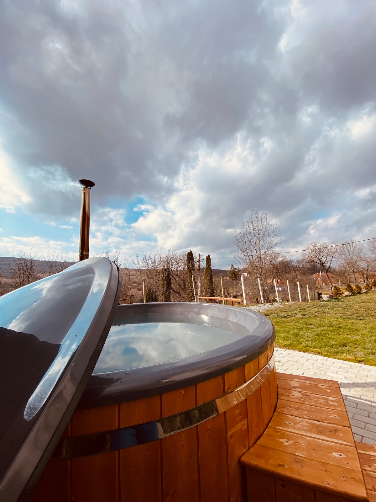 A wooden hot tub is positioned on a wooden deck, with a charcoal stove visible beside it. The surrounding landscape includes grassy areas and trees, under a partly cloudy sky that provides a serene backdrop.