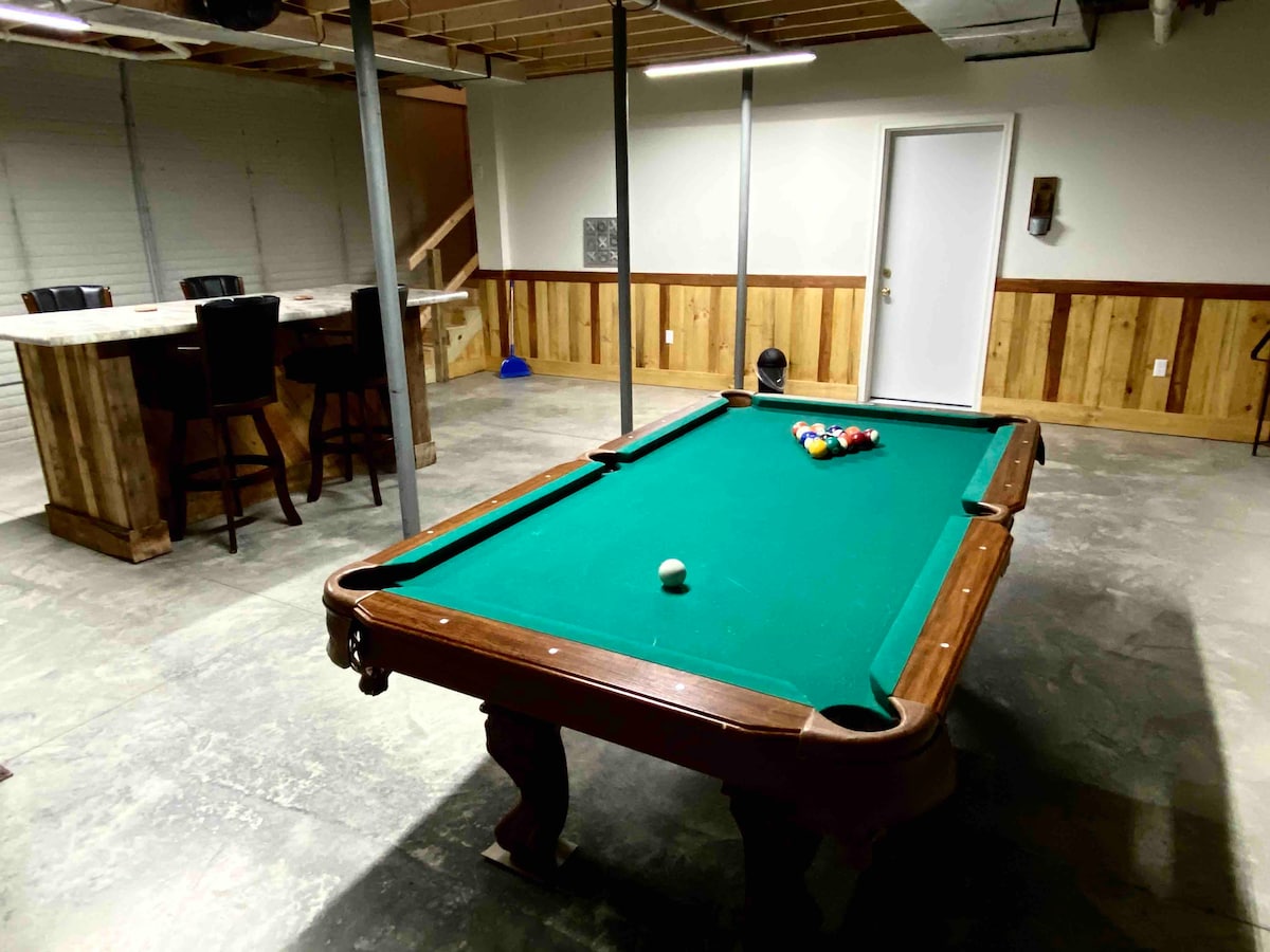An eight-foot pool table occupies the lower level of the home, surrounded by a spacious area featuring wooden paneling. A bar with four stools is visible in the background, adding to the entertainment options available.