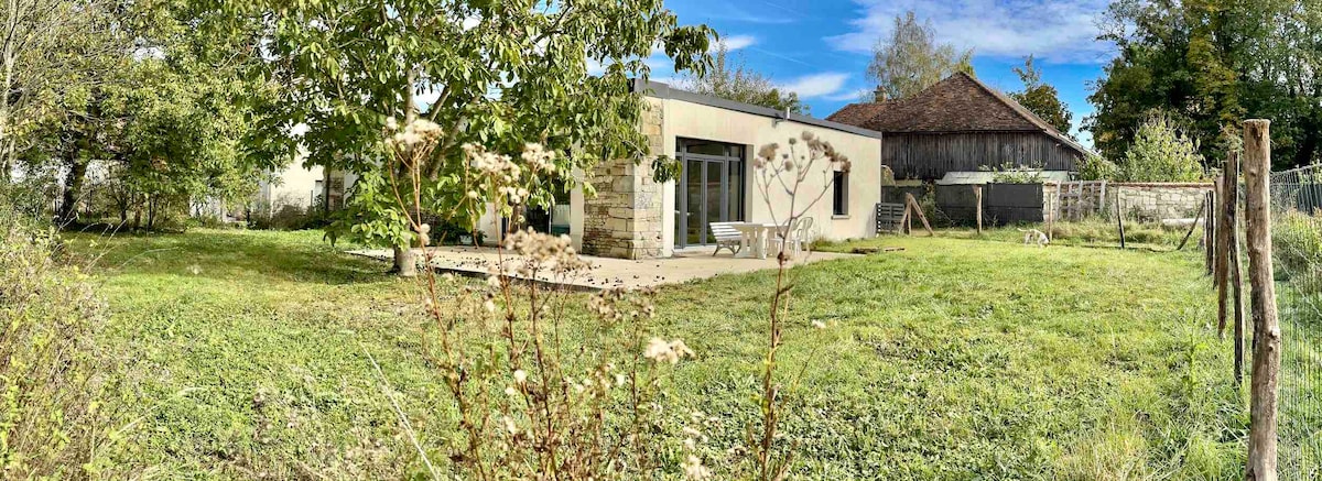 The exterior view captures a modern stone and wood-clad house, surrounded by a lush green lawn and trees. A spacious terrace is visible, providing outdoor access, with the structure's large glass doors offering a glimpse of the inviting interior space.