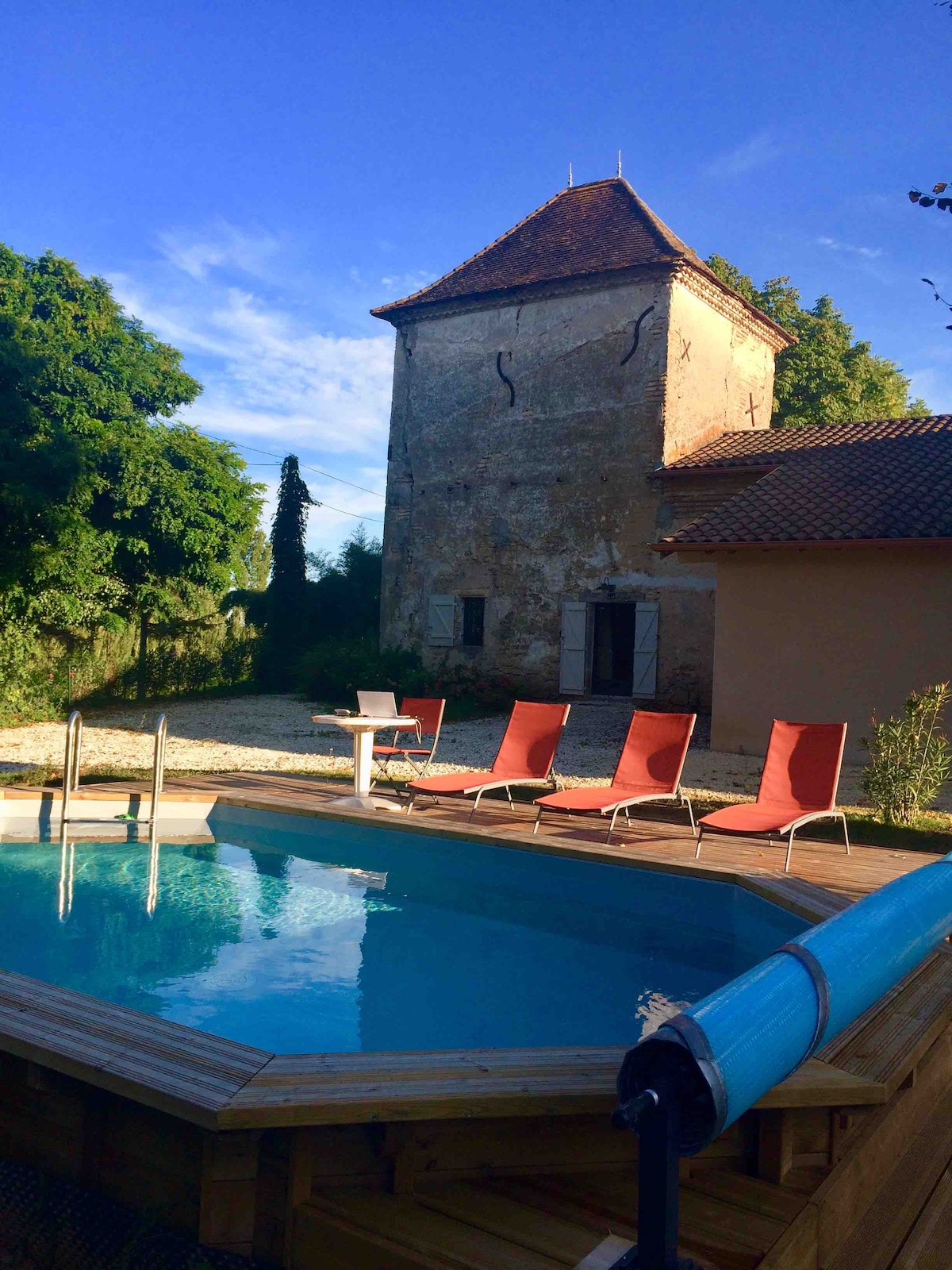 A semi-inground pool is framed by a wooden deck and six lounge chairs in warm tones. Behind, a historic stone building with a distinctive tower is set against a clear blue sky, surrounded by greenery and a gravel area.