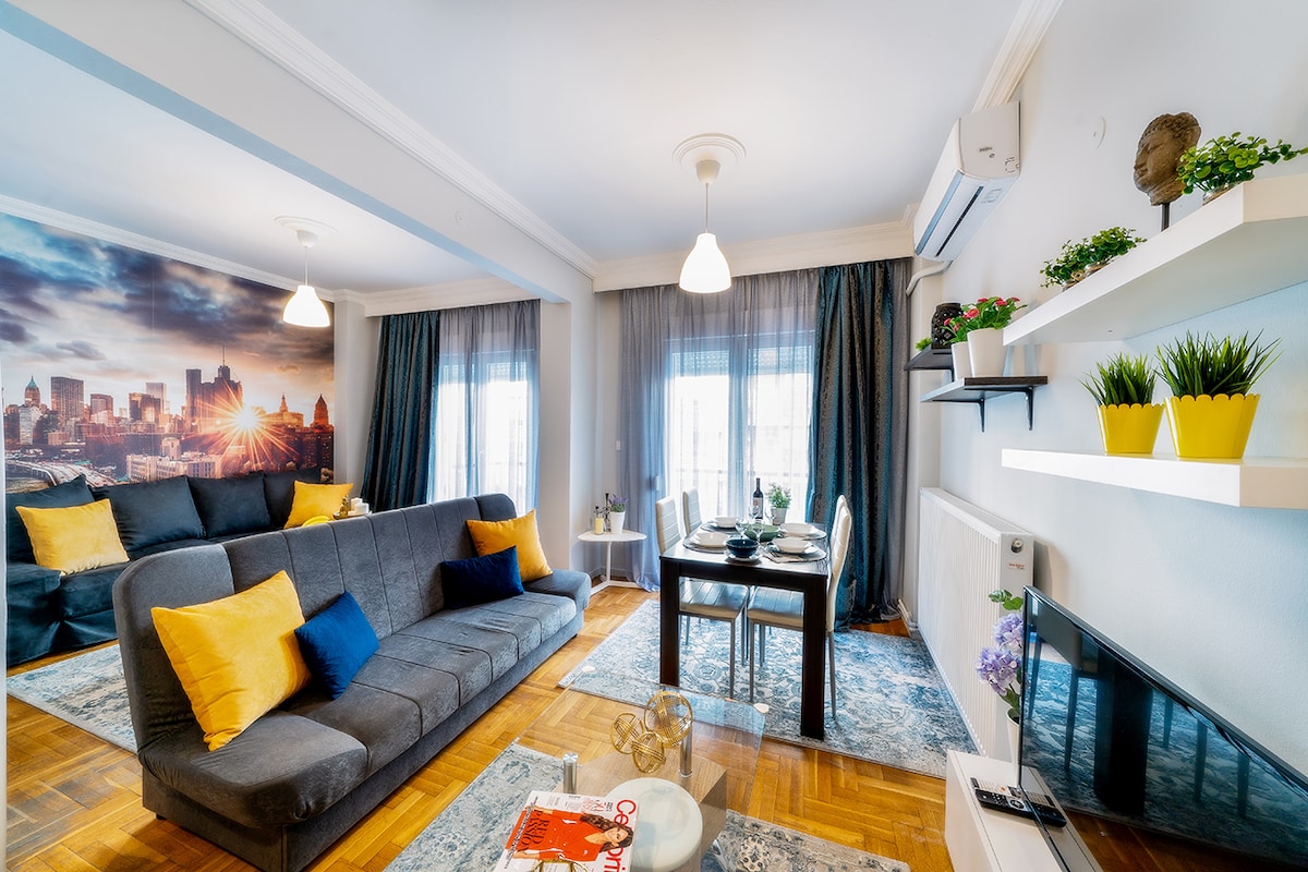 A bright living area is displayed with a comfortable gray sofa accented by yellow pillows. A small dining table with chairs is positioned nearby, set for two. Light-colored rugs lie on the polished wooden floor, and large windows are draped with sheer curtains, allowing natural light to fill the space.