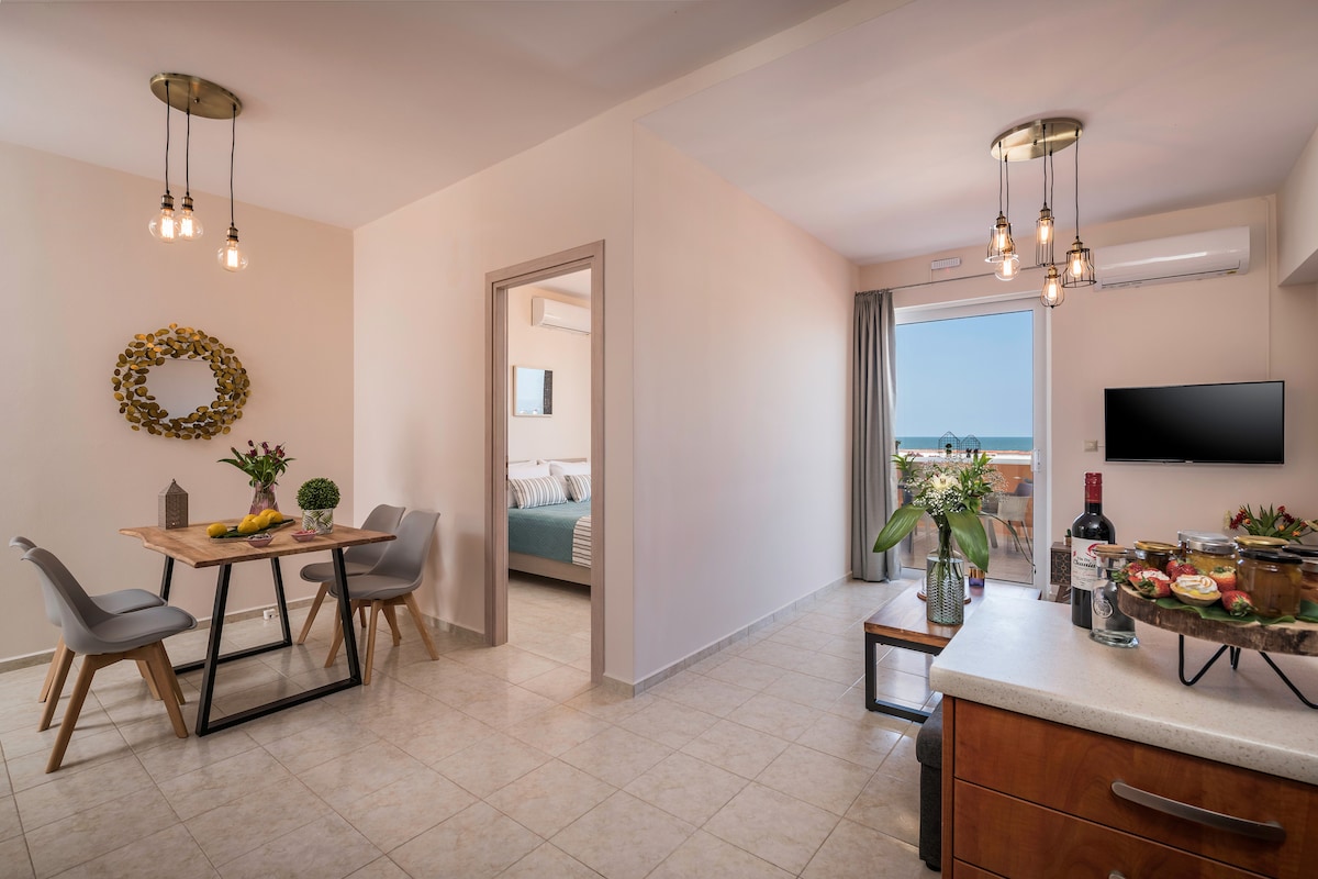 An open living space features a dining area with a wooden table and four chairs. A well-equipped kitchen area is visible, with a variety of fresh fruits displayed. Large windows provide natural light and an unobstructed view of the sea.
