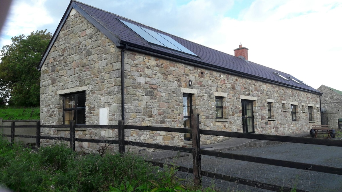 A stone house built from local materials is shown, featuring large windows and a slate roof. The rustic exterior showcases a blend of natural colors, and solar panels are visible on the roof. A wooden fence and gravel pathway lead to the entrance.