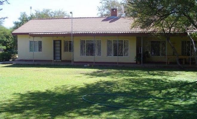 The exterior of the property is showcased, featuring a single-story structure with a tiled roof. Large windows with metal grilles allow natural light into the interior. A well-maintained lawn surrounds the house, offering a serene outdoor space beneath the shade of nearby trees.