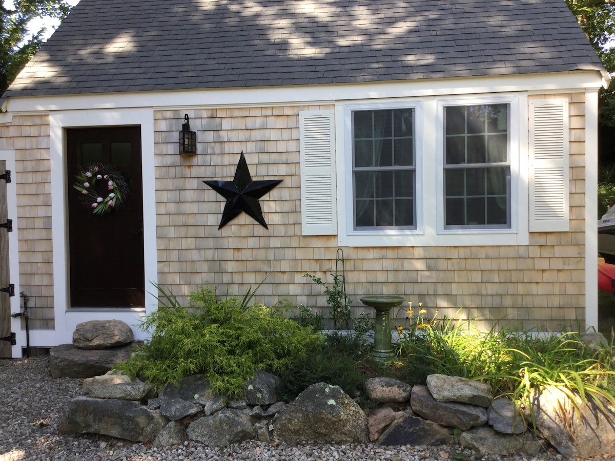 The exterior of a charming cottage features wooden shingle siding and a dark front door adorned with a decorative wreath. A prominent black star decoration is positioned above a pair of windows. Lush greenery and decorative stones create an inviting landscape along the front.