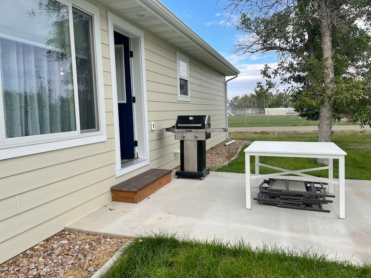 A patio features a propane grill next to a white outdoor table, with several chairs stacked underneath it. The entrance to the home is visible, framed by a large window on one side and a grassy area leading to an open space.