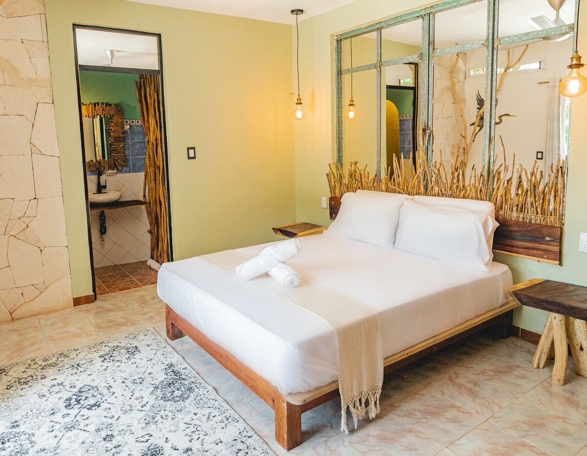 The private room features a spacious bed with fresh linens, paired with wooden bedside tables. A green accent wall complements the natural decor, while soft lighting enhances the ambiance. A mirror reflects the adjacent bathroom area, showcasing neutral tiles and decor elements.