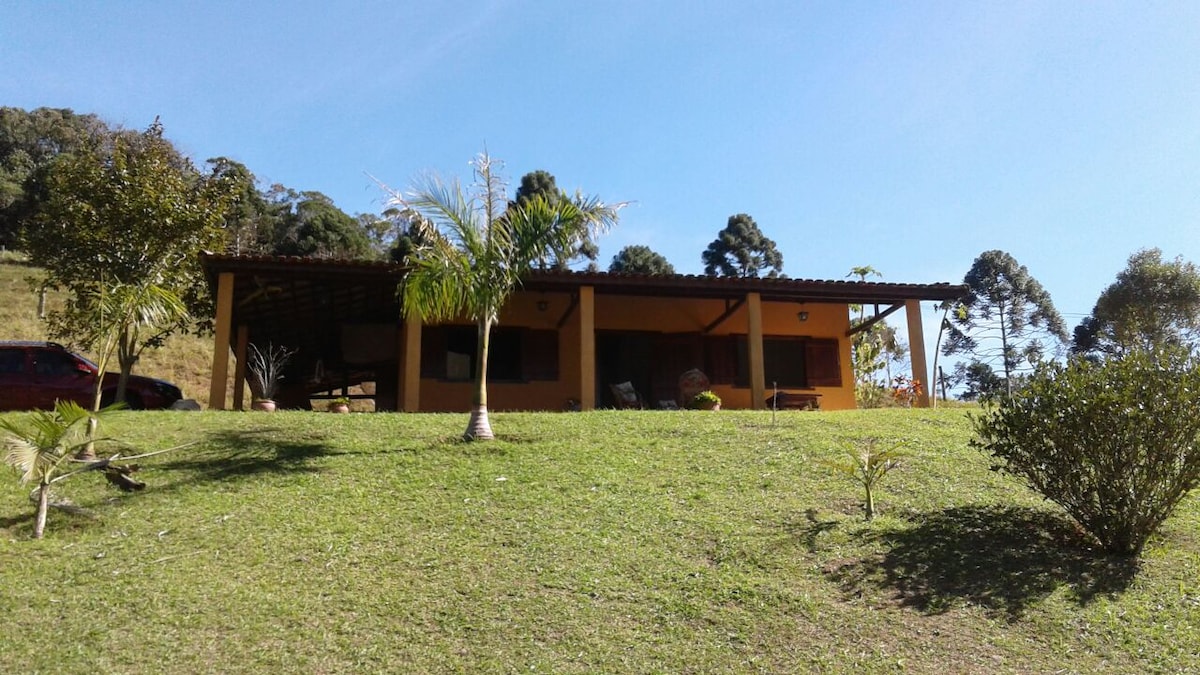 A charming yellow house is set on a grassy hill, surrounded by trees and shrubs. The spacious front porch is shaded, featuring seating areas. The clear blue sky creates a bright and inviting atmosphere, enhancing the natural beauty of the location.