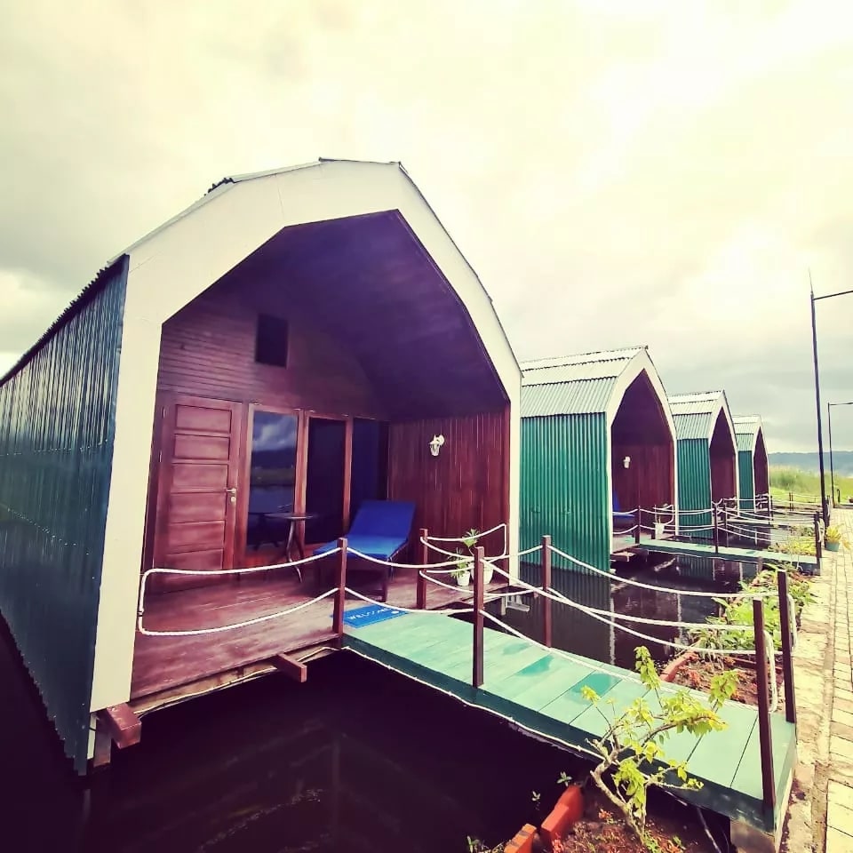 A series of charming cottages are positioned on stilts over water, featuring sloped roofs and wooden accents. Each cottage has a private entrance with a blue chair on the terrace, offering a serene view of the surrounding area.