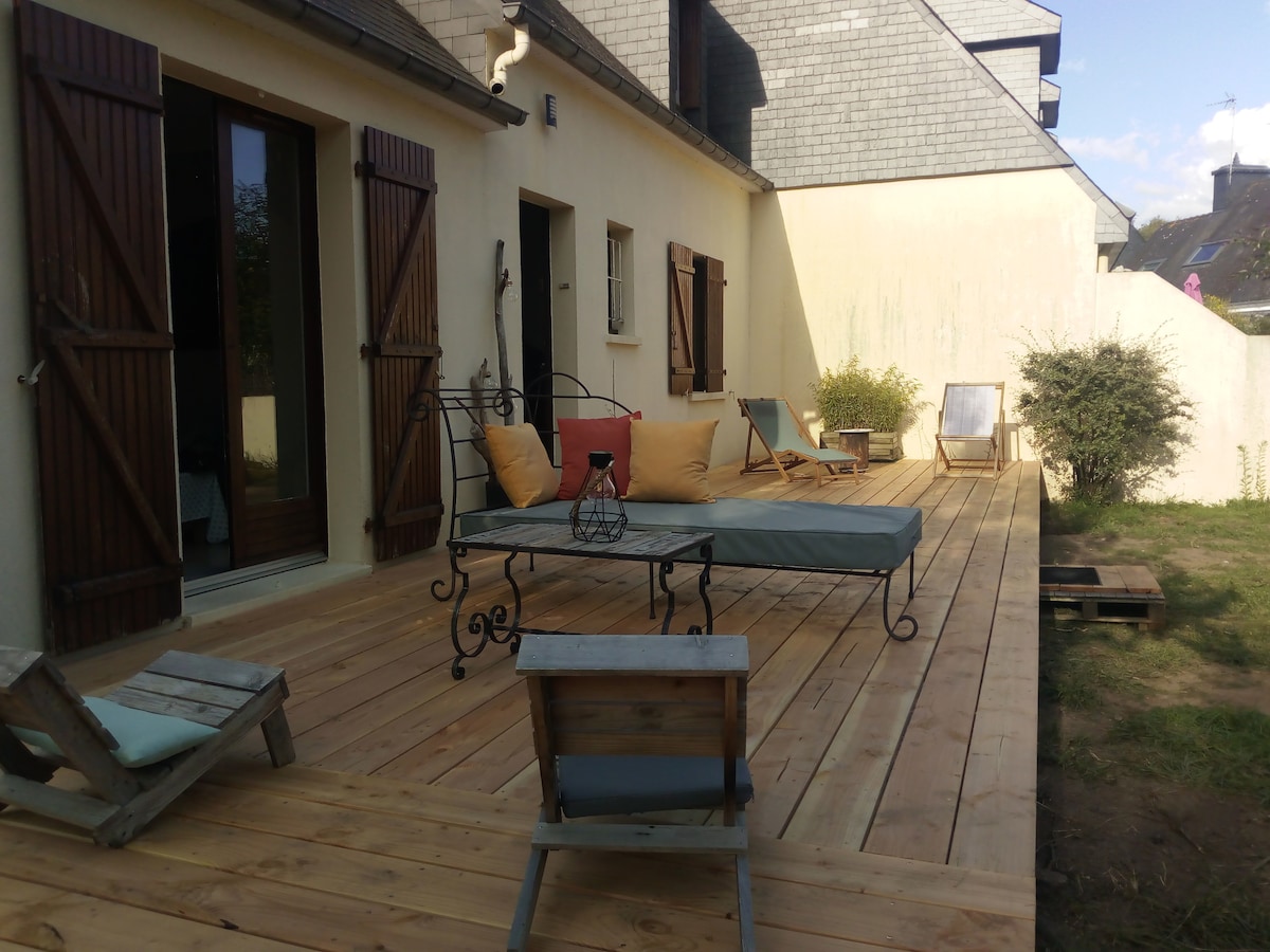 A wooden terrace is depicted, featuring a lounge chair with cushions, a small table, and two additional chairs. The terrace is surrounded by walls, with potted plants visible in the background, offering a private space for relaxation.