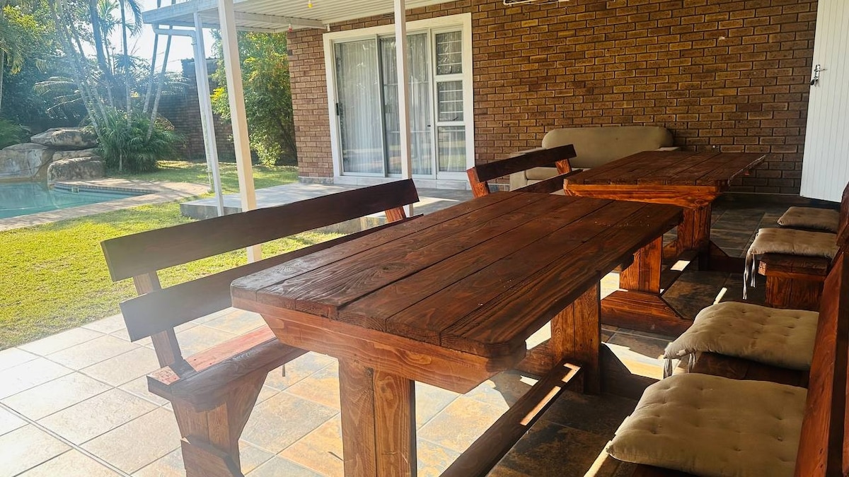 An outdoor dining area features multiple wooden tables accompanied by benches. Cushions are placed on the seating for comfort. The space is shaded, with a view of a grassy yard and a swimming pool in the background, creating a relaxed atmosphere.