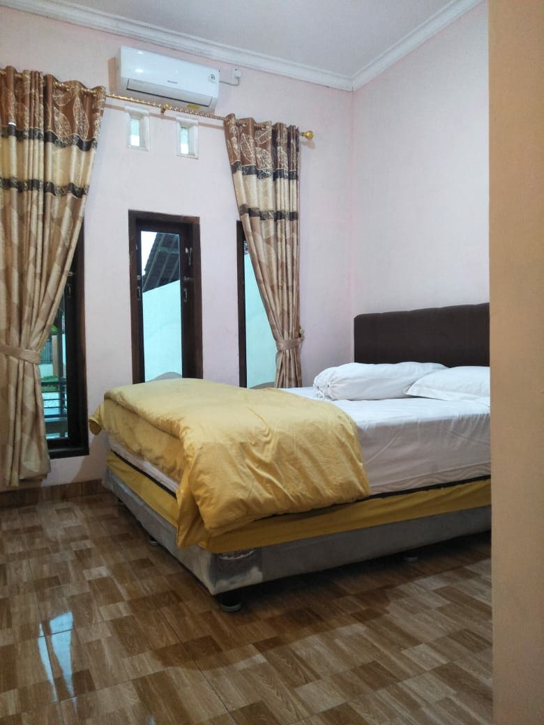 A spacious sleeping area is presented with a bed featuring a light yellow comforter. Two windows allow natural light to enter, with elegant curtains framing them. A wall-mounted air conditioning unit offers comfort, complementing the tiled floor in neutral tones.