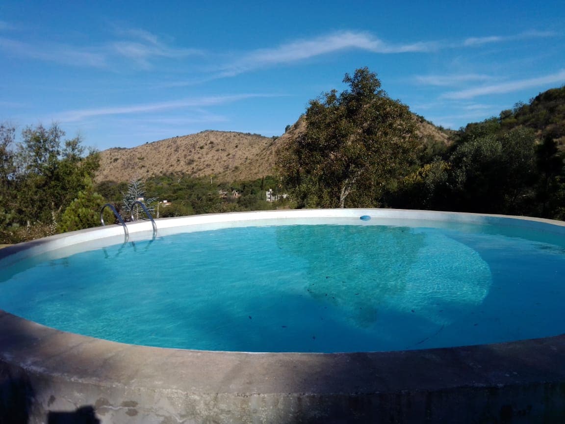 A circular swimming pool is set within a natural landscape, featuring clear blue water. Surrounding vegetation includes trees and hills, creating a serene view against a clear sky. The pool's edge is made of a smooth stone surface, complementing the tranquil environment.