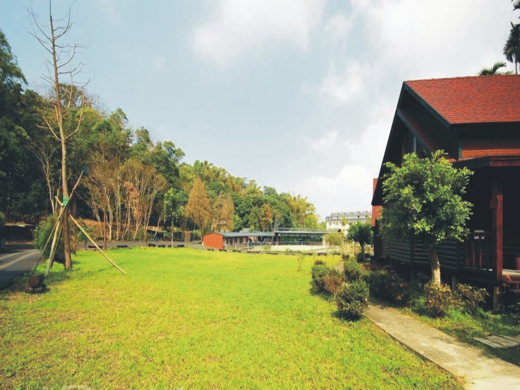 A well-maintained outdoor area is visible, featuring a spacious green lawn surrounded by trees. A pathway leads towards a cluster of wooden structures. Sunlight filters through the canopy, creating a serene atmosphere in this natural setting.