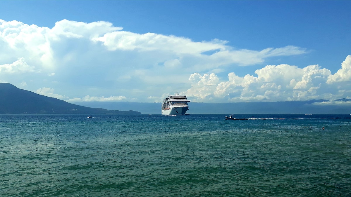 A large cruise ship is anchored in the turquoise water, with scenic mountains visible in the background. The sky is filled with fluffy clouds, creating a tranquil atmosphere. The calm sea reflects varying shades of blue and green, offering a serene coastal view.