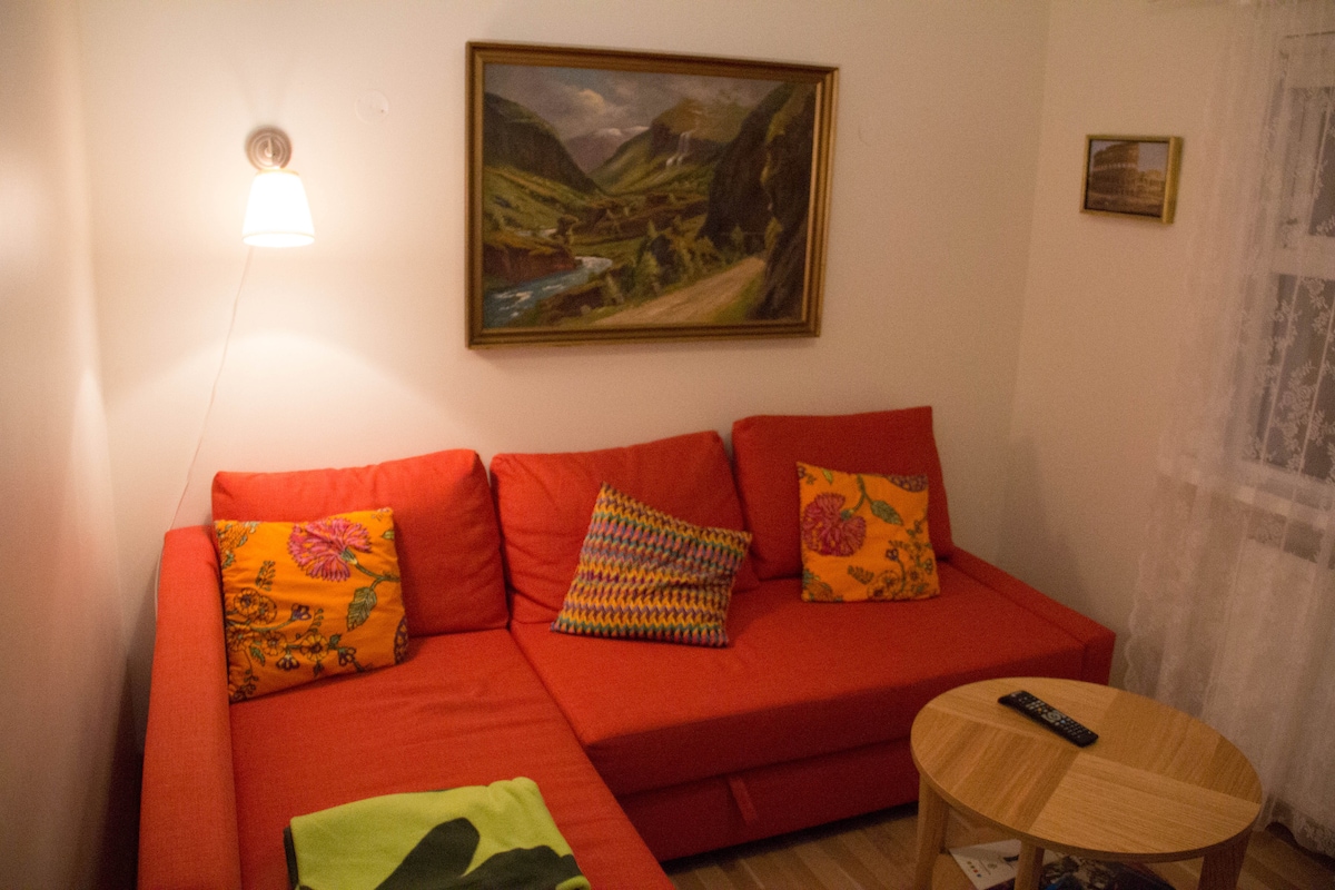 A vibrant orange sectional sofa is adorned with colorful patterned throw pillows. A small round table sits nearby, complementing the seating area. A framed landscape painting adds a touch of nature, while soft lighting enhances the cozy ambiance.