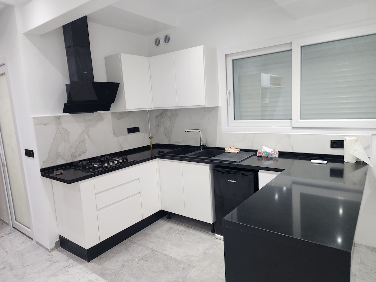 A modern kitchen is showcased with sleek white cabinetry and black countertops. Stainless steel appliances, including a gas stove and refrigerator, are present. Natural light enters through the window, illuminating the clean lines and minimalist design of the space.