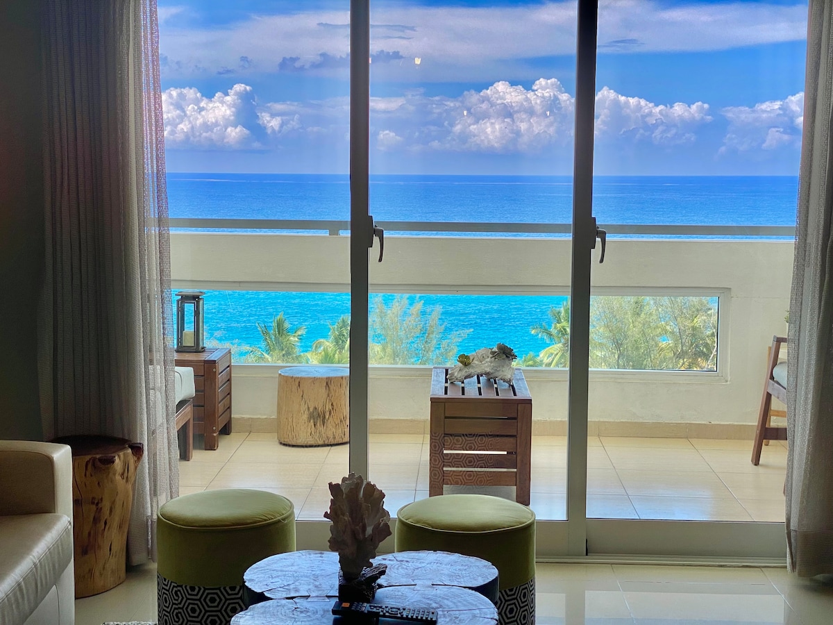 A spacious balcony offers a breathtaking view of the Caribbean Sea, with vibrant blue waters and distant clouds. Wooden furniture pieces provide a natural contrast, while sheer curtains soften the indoor connection to the outdoor scenery.
