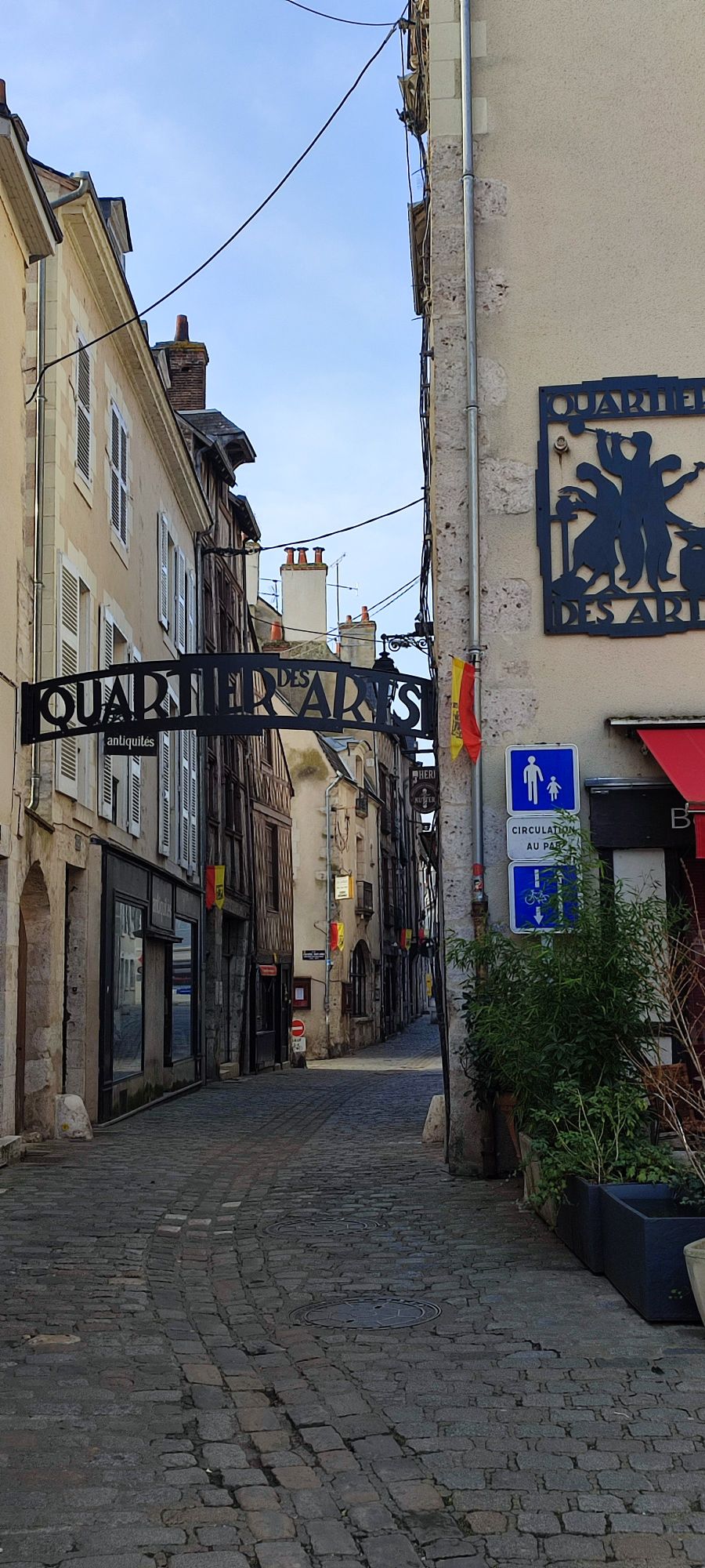 A narrow cobblestone street is seen, featuring buildings with historical architecture and colorful signage. The 'Quartier des Arts' archway stands prominently overhead, welcoming visitors. Potted plants are positioned along the sidewalk, adding a touch of greenery to the charming urban setting.