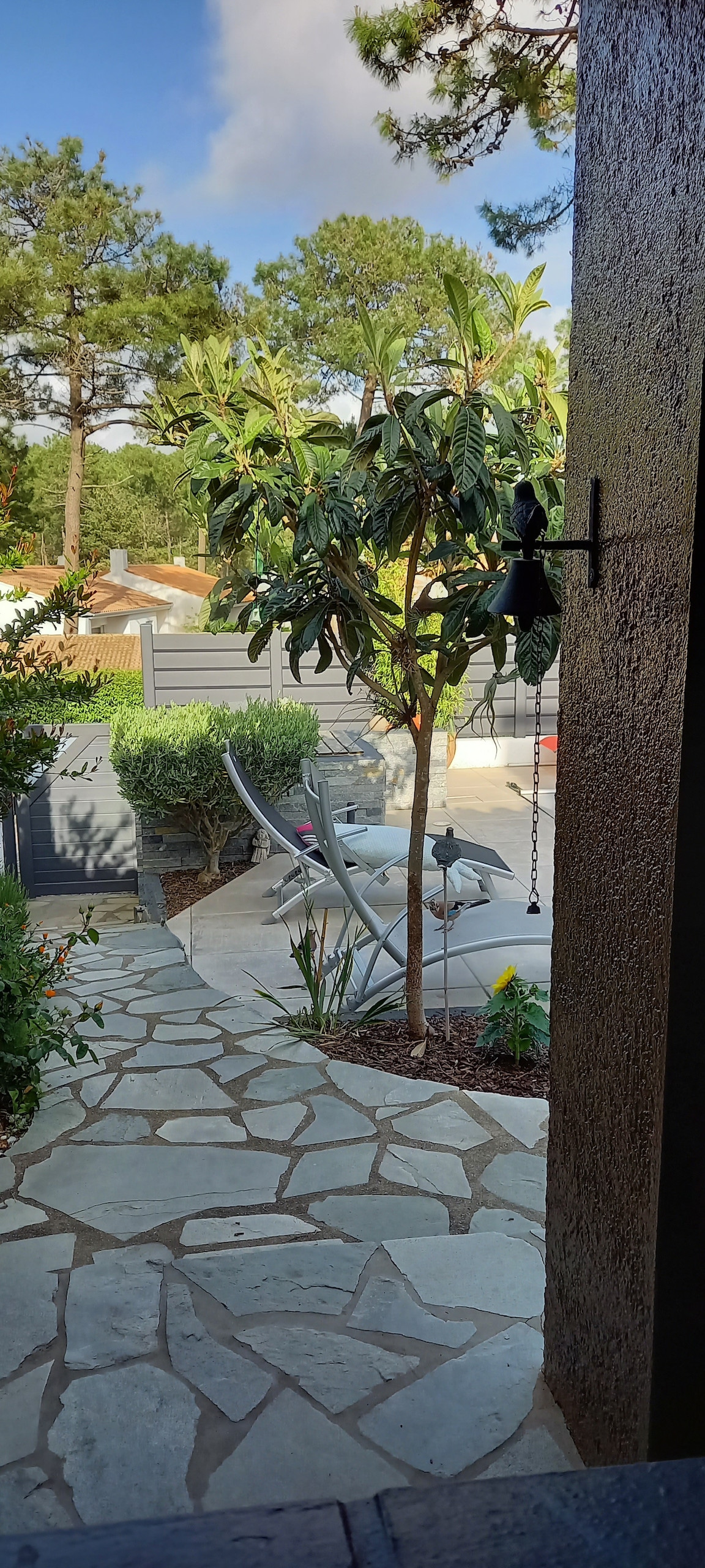 A paved stone path leads through a lush garden towards a serene outdoor area with a pool. A tree provides shade, while lounge chairs are visible nearby, creating a relaxed atmosphere. The view frames a sunny landscape, emphasizing the peaceful surroundings.