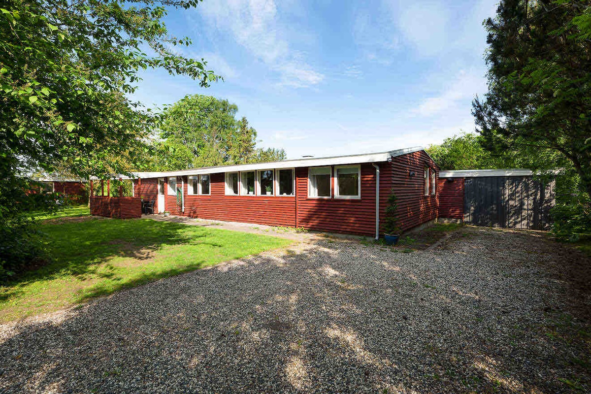 A single-story red building is framed by greenery, featuring multiple windows that invite natural light. A gravel pathway leads to the entrance, providing clear access. The surrounding area is predominantly grass with a few trees, creating a serene outdoor environment.