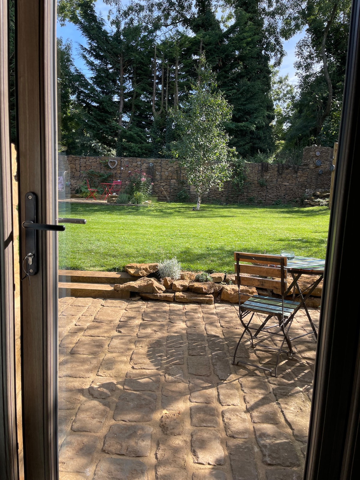 A private garden is visible from an open door, featuring a well-maintained grassy area surrounded by trees. A small table and chair set rests on a stone patio, with natural stone borders enhancing the outdoor space.