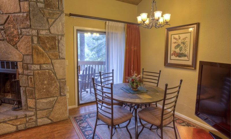 A dining area features a round table surrounded by six chairs, with a decorative centerpiece of flowers. A large window with sheer curtains offers views of the outdoors, while a stone fireplace adds warmth to the space. A chandelier hangs overhead, providing ambient lighting.