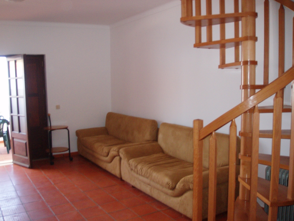 A comfortable living area features a tan fabric sofa positioned against a wall. A wooden spiral staircase leads to the upper floor. The space is illuminated by natural light from a nearby door, which opens to an outdoor area.