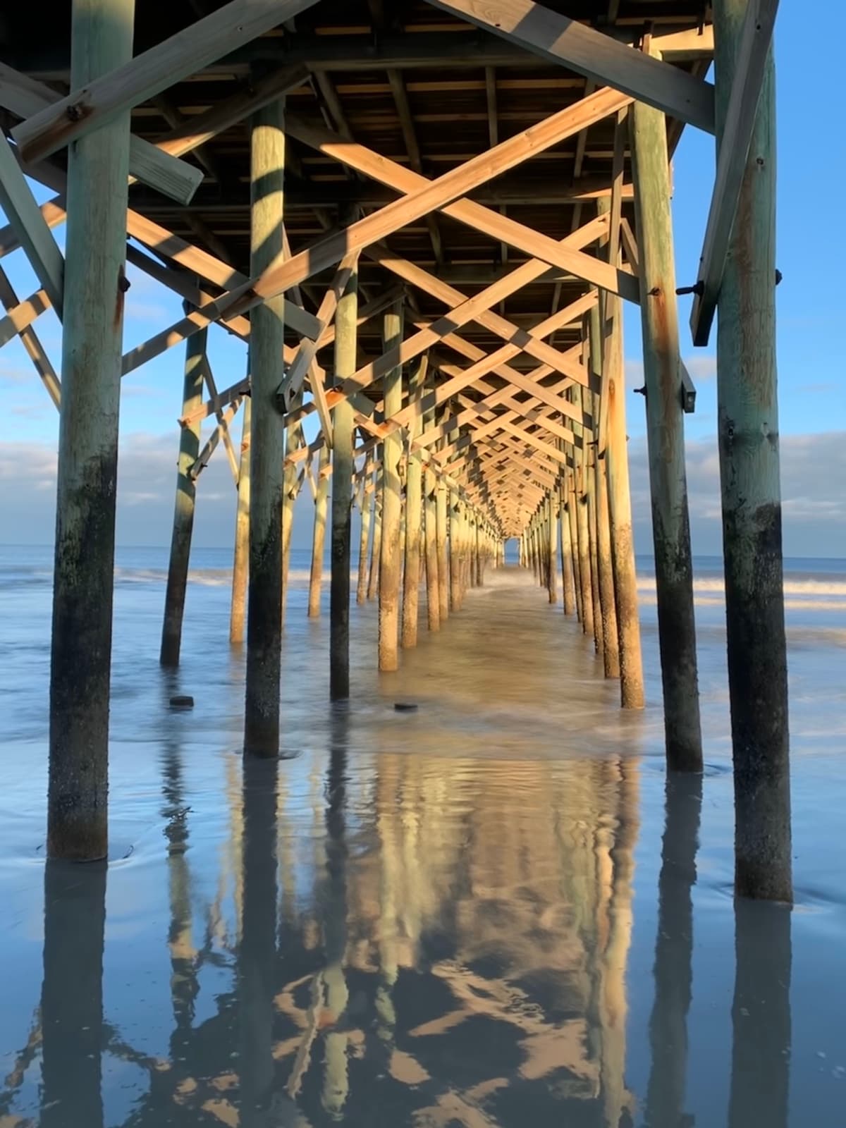 A wooden pier extends into the water, supported by tall columns. Reflections of the structure are visible on the wet sand, creating a serene atmosphere. The sky showcases soft clouds, and gentle waves lap at the edge of the beach, indicating a tranquil seaside environment.