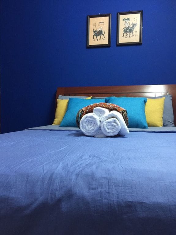 A sleeping area features a spacious bed with a blue blanket and decorative pillows in vibrant shades of blue and yellow. Rolled towels are neatly arranged on the bed, complementing framed artwork on the wall in the background.