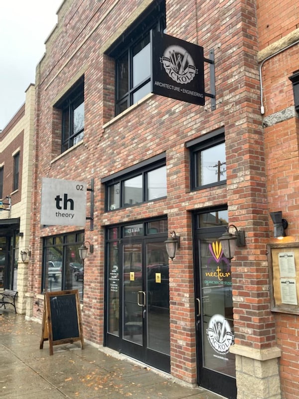 A brick facade is seen with large glass doors and windows. Signage above denotes different businesses, including 'theory' and 'NECTAR,' designed in modern typography. Outdoor seating is visible nearby, enhancing the streetscape of downtown Bozeman.