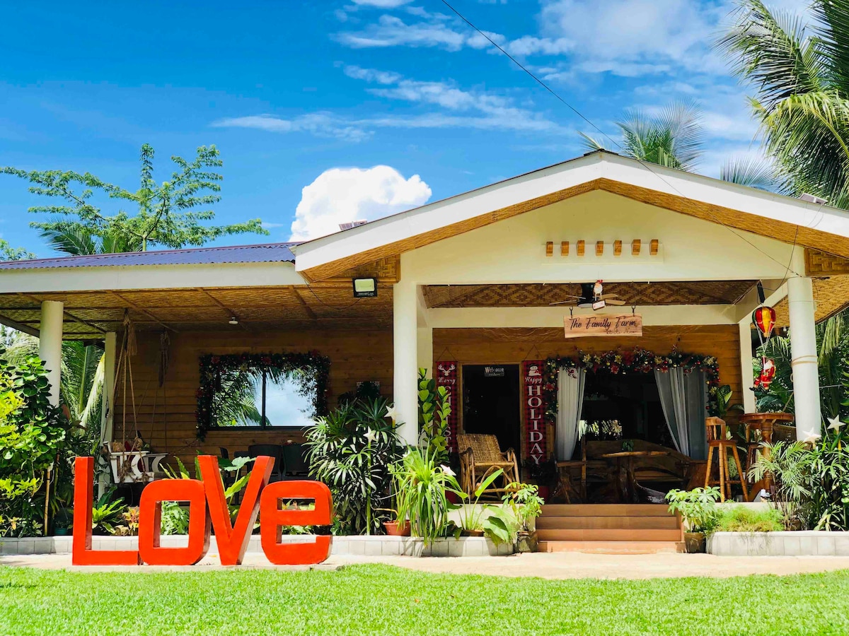 A welcoming exterior of a vacation home is surrounded by lush greenery and palm trees. The structure features a large, covered porch adorned with festive decor. A prominent red sign spelling 'Love' is displayed in the front yard, creating an inviting ambiance.
