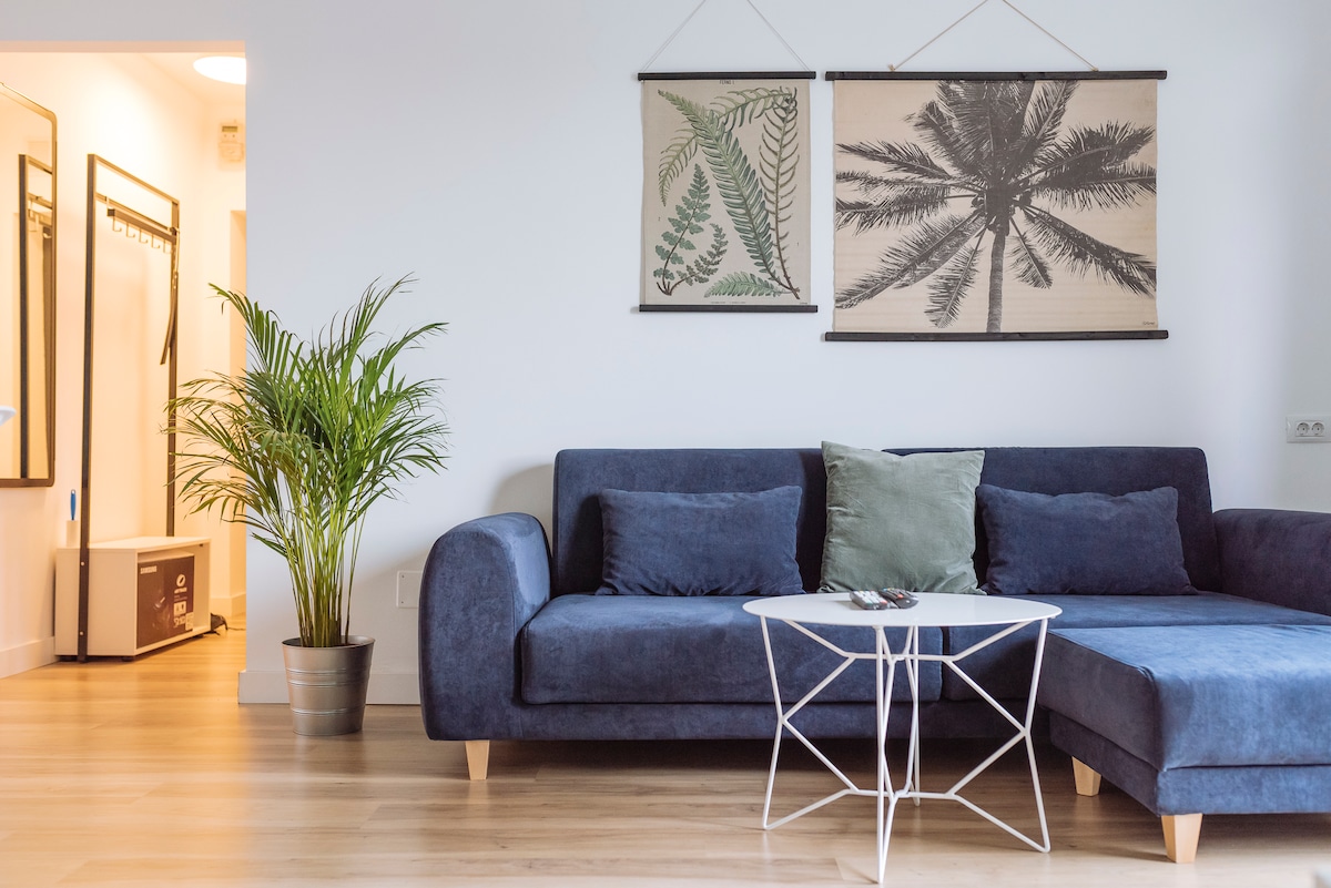 A spacious living area is highlighted by a modern blue sofa with decorative pillows. A white metal coffee table sits in front, complemented by framed tropical art on the wall. Natural light enhances the tranquil atmosphere, and a potted plant adds a touch of greenery.