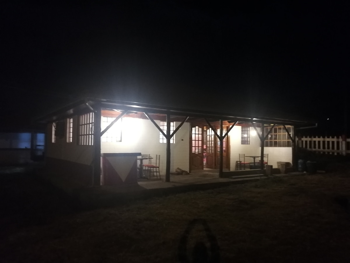 The exterior of the unique country house is illuminated by soft outdoor lighting at night. The structure features a welcoming porch with two tables and chairs, allowing for outdoor seating. Large windows reflect a warm glow from the interior, inviting guests to enjoy the tranquility.