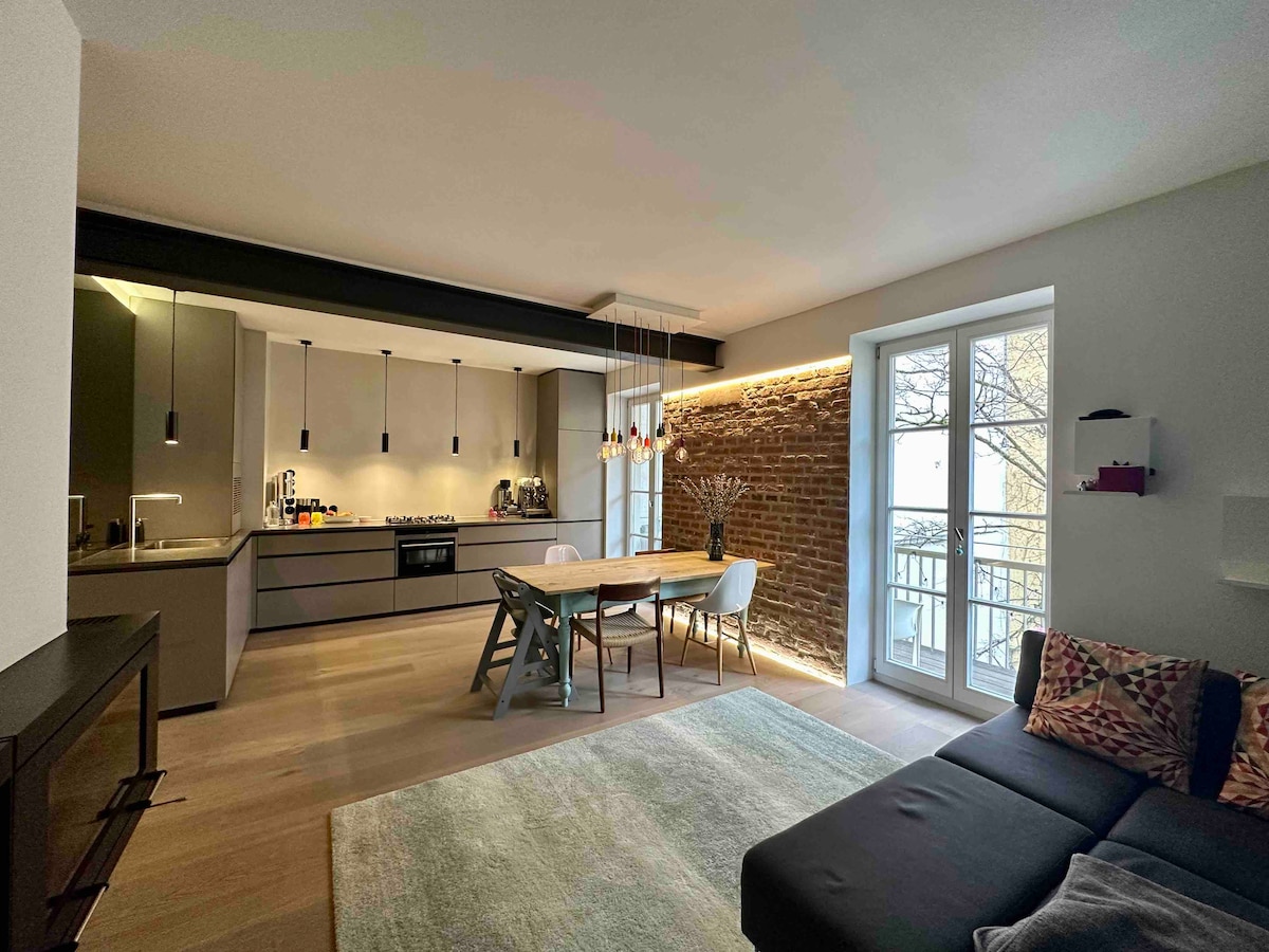 A modern kitchen area features sleek cabinetry and high-end appliances, complemented by a dining table surrounded by chairs. Exposed brick adds character to the space, while large windows allow natural light to illuminate the room. A comfortable sofa is positioned nearby with soft textiles.