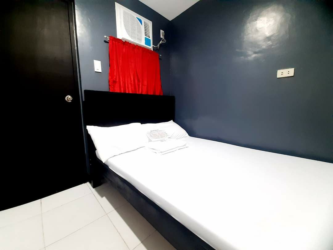 The room features a minimalist design, highlighting a spacious bed with fresh linens. A red curtain frames a window, and a wall-mounted air conditioning unit is present. The smooth, dark-colored walls contrast with the white tiled floor, creating a modern aesthetic.