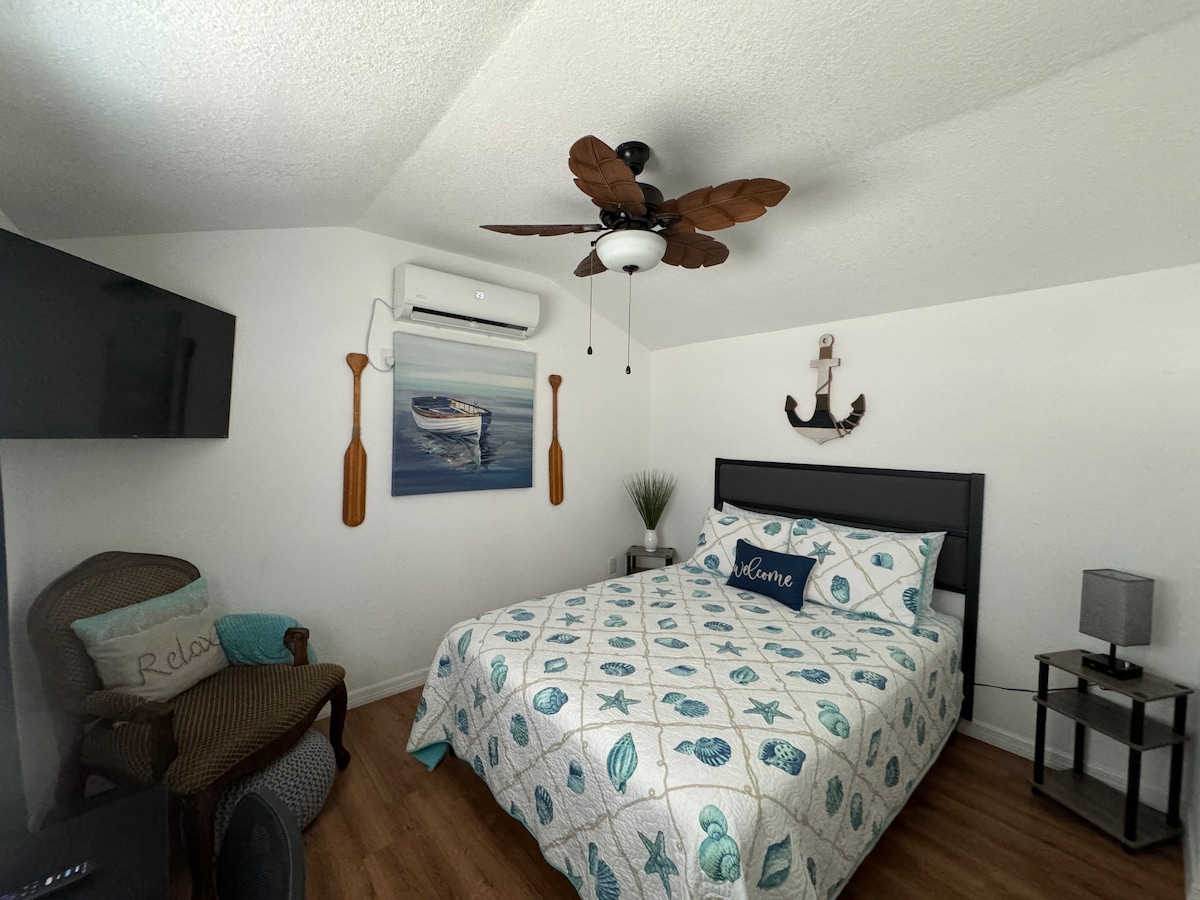 A cozy bedroom features a spacious bed adorned with a sea-themed quilt and decorative pillows. A ceiling fan and wall-mounted air conditioning unit provide climate control. Wall art depicting nautical elements and a decorative anchor add character. A small nightstand and a comfortable chair complete the space.