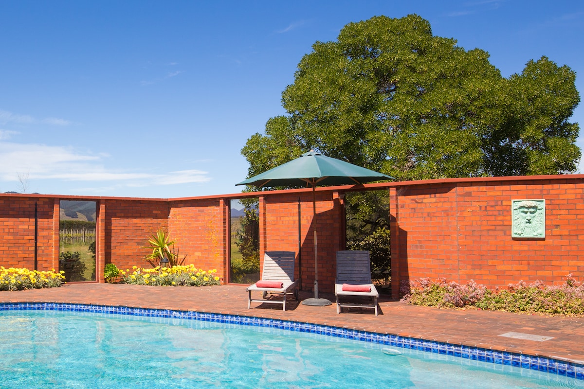 A refreshing pool area is surrounded by a brick wall, offering privacy. Two lounge chairs with a green umbrella are positioned nearby, complemented by colorful landscaping. A tree is visible in the background, providing shade and enhancing the outdoor setting.